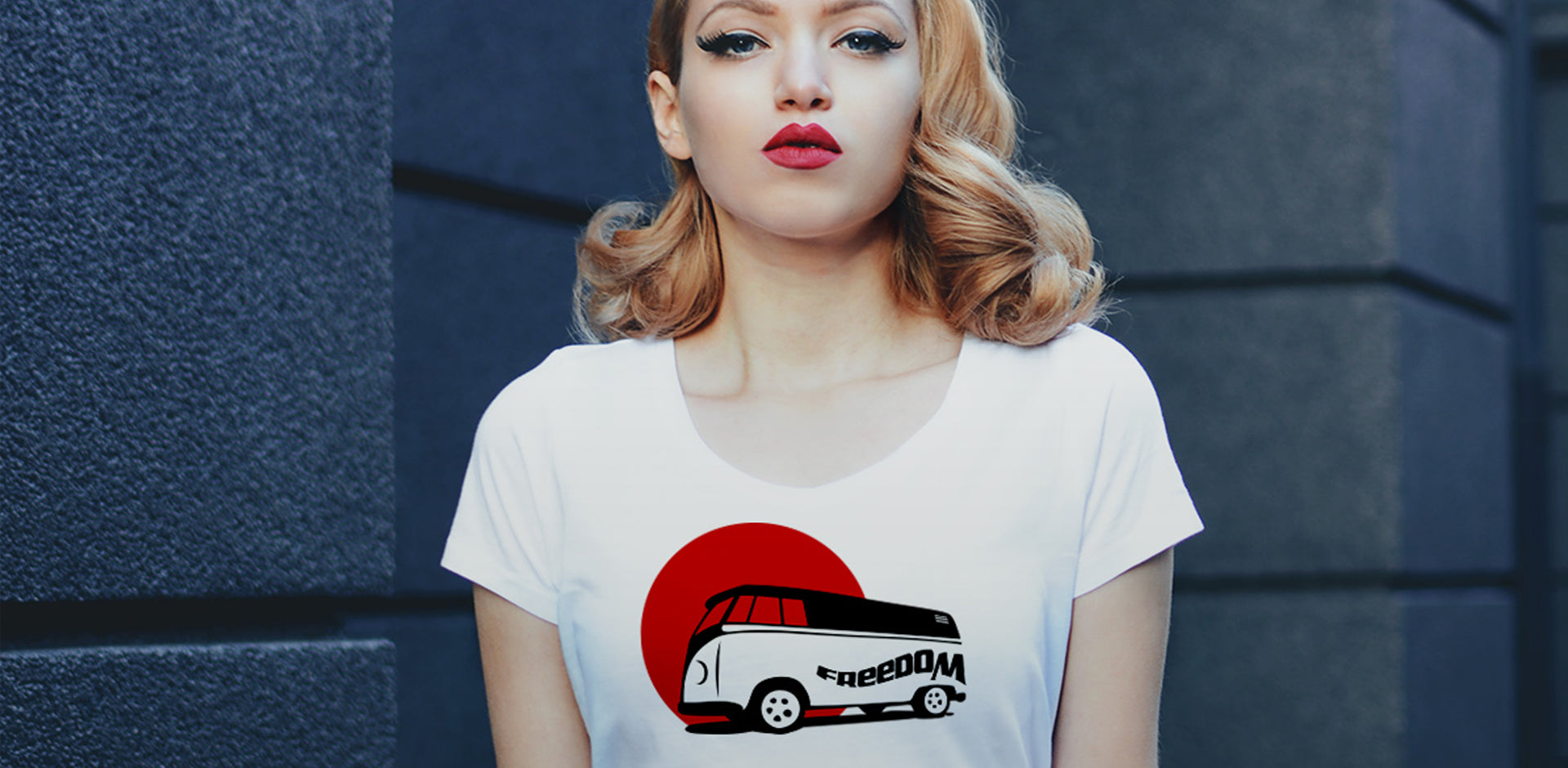 Eine Frau mit hellen, lockigen Haaren und rotem Lippenstift trägt ein weißes T-Shirt. Auf dem Shirt ist ein VW Bus im Stil der 70er Jahre abgebildet, der vor einem roten Kreis steht, der an eine aufgehende Sonne erinnert. Das Wort "Freedom" ist in schwarzer Schrift auf der Seite des Busses zu sehen.